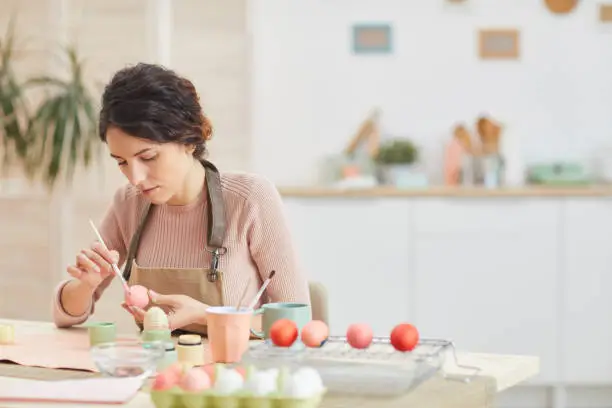 Portrait of adult woman painting eggs in pastel colors for Easter while sitting at table in cozy kitchen interior, copy space