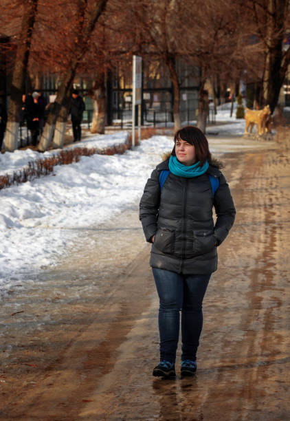 A young girl in a black jacket and jeans walks along the path in a city park on a sunny spring day. The warm sun melted the snow on the tracks. She enjoys the warm weather. Early spring in the city.