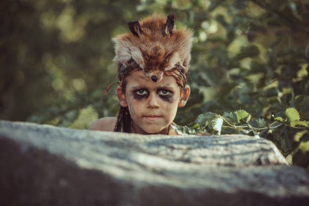 Caveman, manly boy hunting outdoors. Prehistoric tribal boy outdoors on nature. Young shaggy and dirty savage, warrior and hunter hiding in an ambush behind a stone. Primitive ice age man in animal skin portrait. Actor playing role in movie. Reconstruction of Neanderthal and cro-magnon life