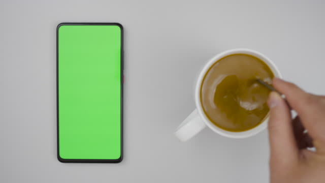 Man Hand Cup of Hot Coffee and Using Smartphone Watching Green Screen Top View. Smartphone with Green Mock-up Screen Business Concept. Person Hand Stirring Coffee with Spoon on Table. Slow Motion.