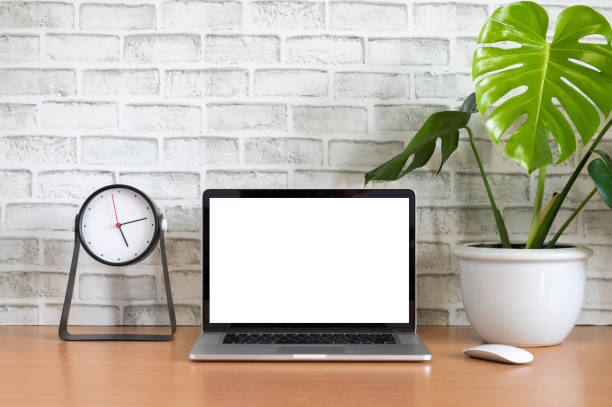 Blank screen of Laptop computer with clock, tablet, mobile phone, mouse and monstera plant pot on wooden table, White brick background