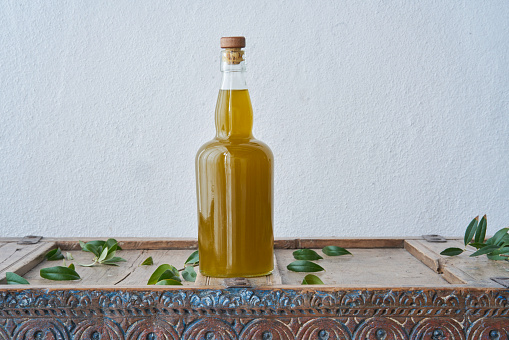 Still-life of glass bottle on a wooden rustic piece of furniture filled with fresh olive oil surrounded by green olive leaves against a white wall in a country house