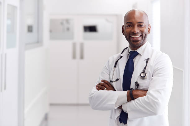portrait of mature male doctor wearing white coat standing in hospital corridor - doutor imagens e fotografias de stock