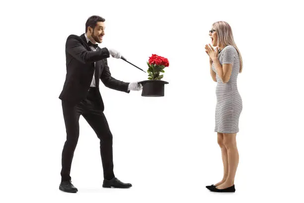 Full length shot of a magician making a magic trick with flowers and a hat in front of a surprised young woman isolated on white background