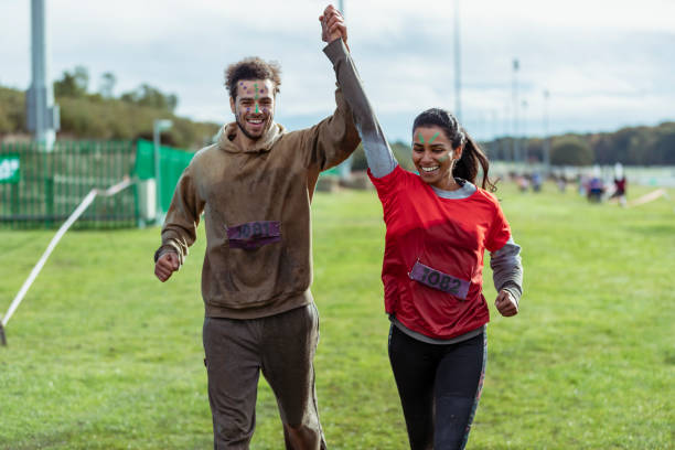 Mixed race couple finishing Stampede race together Mid adult couple finishing charity run, holding hands up in the air, relief, fun, achievement resilience photos stock pictures, royalty-free photos & images