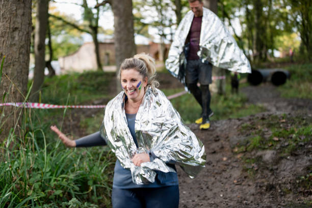 Woman wearing foil blanket running along woodland path Mature charity runner running in countryside for Stampede, fundraising, excitement, fitness people covered in mud stock pictures, royalty-free photos & images