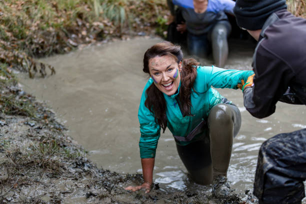 Cheerful woman being helped out of muddy pool Happy mature woman being pulled from dirty pool on cross country run, determination, achievement, success stampeding photos stock pictures, royalty-free photos & images