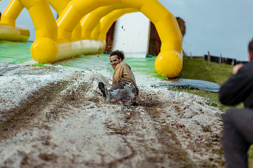 Smiling mid adult man laughing on mudslide by inflatable, dirty, splashing, carefree