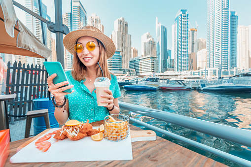 Asian girl has Breakfast with seafood cuisine in a restaurant on the terrace overlooking the skyscrapers and the Marina port in Dubai and communicates with friends on a smartphone
