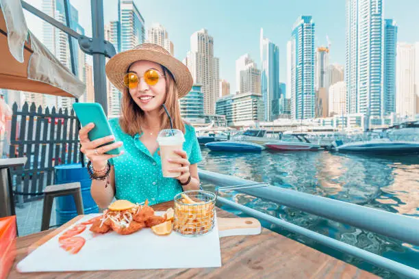 Photo of Asian girl has Breakfast with seafood cuisine in a restaurant on the terrace overlooking the skyscrapers and the Marina port in Dubai and communicates with friends on a smartphone