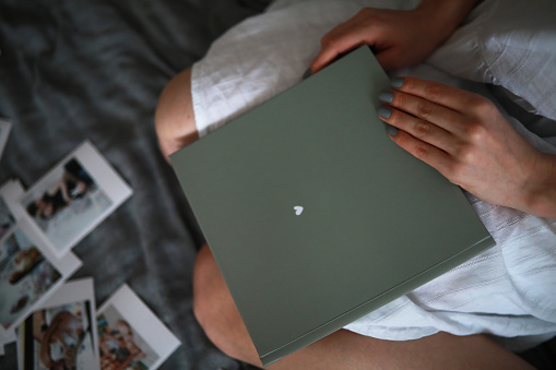 A woman sits on a bed and holds a photo album in her hands on the background of laid out photos. Top view. Closeup.