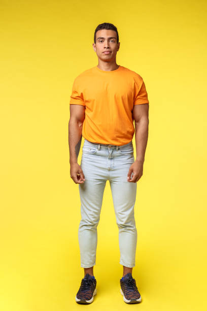istock Full-length vertical shot masculine hipster guy, hispanic ethnicity, standing casually yellow background, wear stylish orange t-shirt, white pants, look camera with no expression, slightly smiling 1204020332