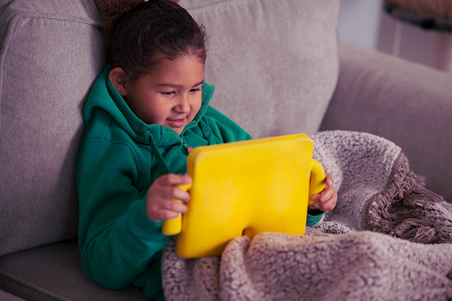 Kids screen time concept - smiling 6 years old, mixed-race girl sitting on the sofa in the living room and watching tablet computer with the yellow case.