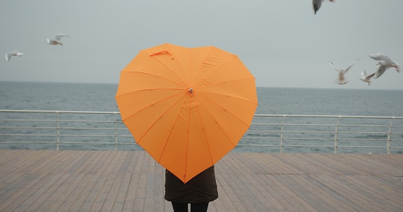 Woman in parka with orange umbrella in heart shape enjoying sea view with seagulls in storm. Overcast weather.