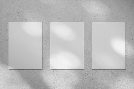 Three empty white vertical rectangle poster or business card mockups with diagonal dappled light spots on gray concrete wall. Flat lay, top view. For advertising, brand design, stationery presentation