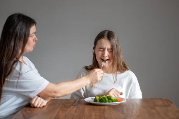 mutter zwingt ihre weinende teenager-tochter, gesunde nahrung zu essen, die ihren brokkoli mit gabel füttert. lebensmittelverschwendung. kein veganes ernährungskonzept. keine wahl - conflict one way sign road sign stock-fotos und bilder