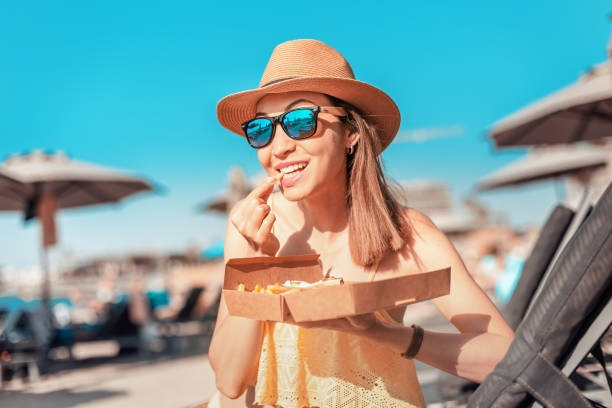 ragazza felice prendere il sole sulla spiaggia e fare spuntini con patatine fritte fast food - heat female summer vacations foto e immagini stock