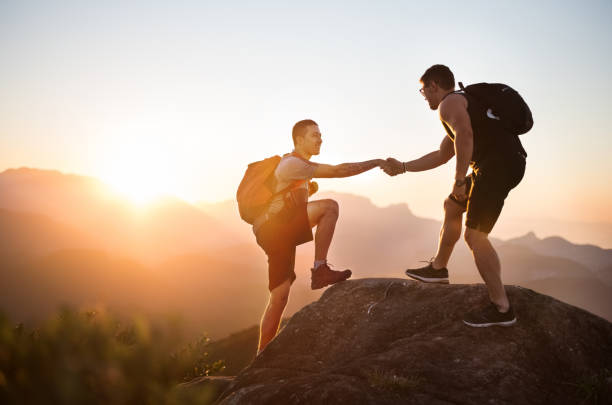 Hiking is a great adventure Shot of a two young men on mountain peak climbing and helping each other climbing up a hill stock pictures, royalty-free photos & images