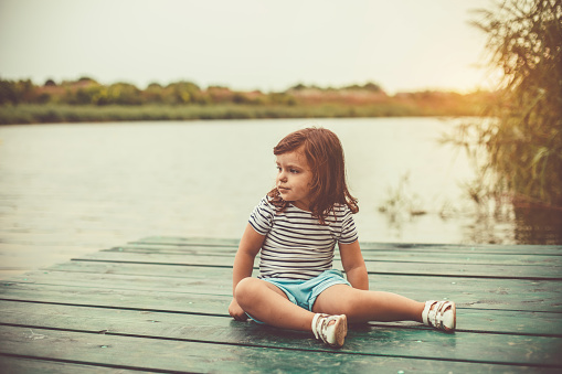 Full length shot of cute little toddler sitting on a pier and looking in the distance at the lake while relaxing and enjoying warm summer day.