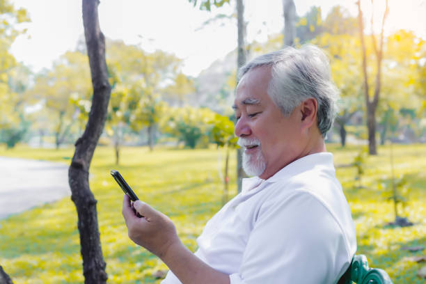 ハンサムなシニアアジアの男性は、公園や高齢者のインターネットオンラインサービスを使用してスマートフォンのビデオ通話で家族とおしゃべりし、ベンチに座って、老人は幸せでニュー� - one senior man only grandfather portrait old ストックフォトと画像
