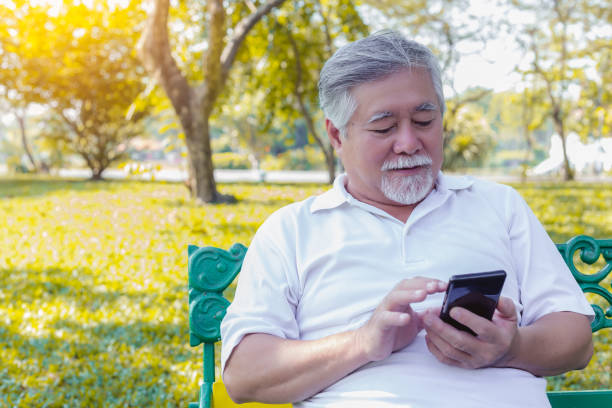 シニアアジアの男性は、公園でスマートフォンやインターネットオンラインを使用して家族とおしゃべり。老人家族は外国に滞在します。老人は彼らとおしゃべりすると幸せになる。祖父は� - one senior man only grandfather portrait old ストックフォトと画像