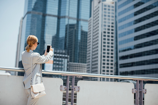 Pretty elegant businesswoman standing at the city by the skyscrapers and taking photo with her cell phone.