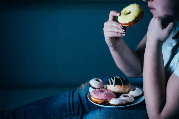 Overweight woman laying on sofa eating sugary food Overweight depressed woman laying on sofa eating sugary food watching TV. Sugar addiction, unhealthy lifestyle, weight gain, dietary, healthcare and medical concept. over eating stock pictures, royalty-free photos & images