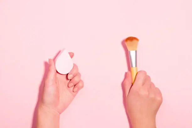 Woman holding makeup brush and beauty blender on pastel pink background. Beauty accessories with makeup brush versus beauty sponge for foundation