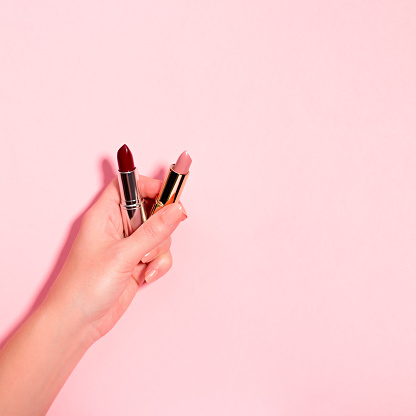 Woman holding dark and light lipsticks on pastel pink background. Day makeup versus night makeup concept. Dark red and light pink, nude lipsticks in woman hands on pastel pink, top view