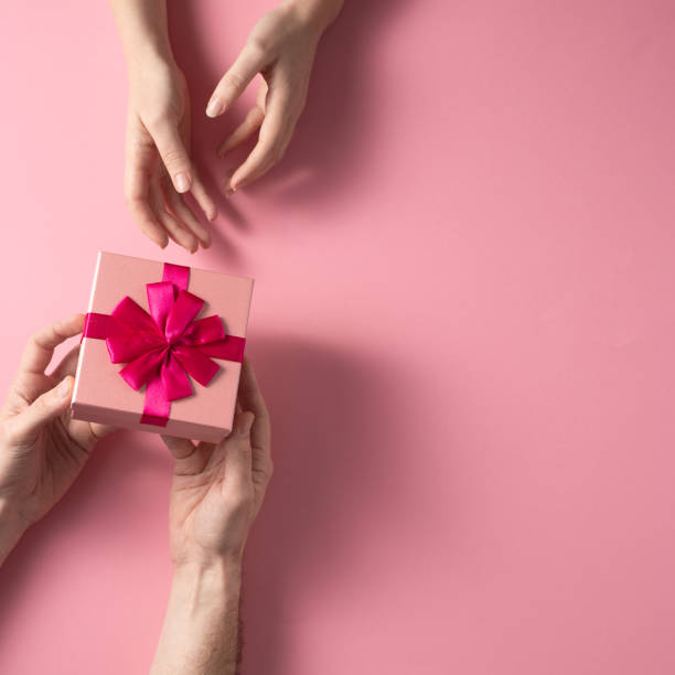 istock Valentine's Day celebration concept. A nice gift from a loved one. Box with a bow hands of a man and a woman on a delicate pink background. Copy space. Flat lay.Square. 1204092879