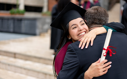 Portrait of a happy student hugging her father on graduation day while holding her diploma