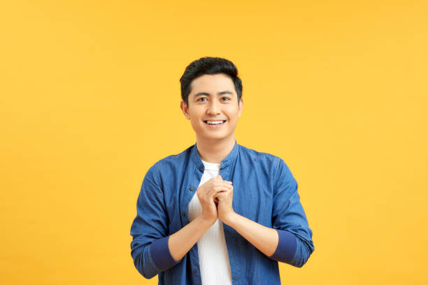 istock Young asian man clasping hands and looking at camera against yellow background 1204101714