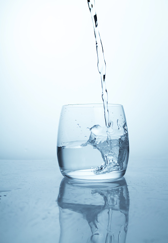 A stream of water flowing into a transparent glass cup with a splash of drops and drops. A jet of clean drinking cold refreshing spring water pouring into a glass