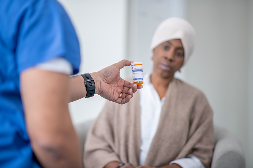 An African woman with cancer sits with her male nurse as he hands her a new prescription and goes over how to take it.  The African woman is dressed comfortably in a white shirt with a light brown shall over her shoulders and a head scarf on.  She has a neutral expression on her face as she listens attentively.  The male nurse has his back to the camera and is wearing blue scrubs.
