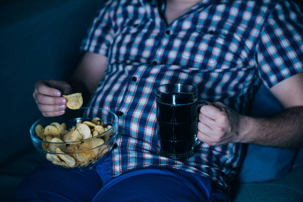 overweight man watching tv with junk food and beer overweight man watching tv with junk food and beer. overeating, sedentary lifestyle, bad habits, food addiction, eating disorders unhealthy living stock pictures, royalty-free photos & images