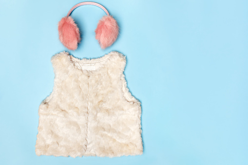 A set of warm clothes. Fluffy pink earmuffs and a white vest on a blue background. Top view flat lay copy space.