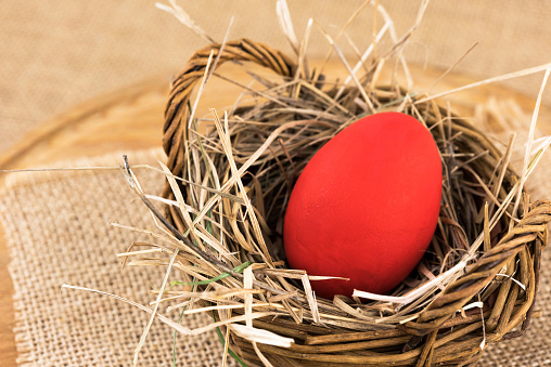 Close up shot of a red Easter egg in woven basket with dry grass against beige background. Egg in a nest concept.