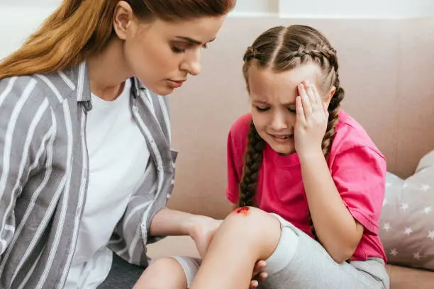 mother holding leg with wound on knee while emotional daughter crying at home
