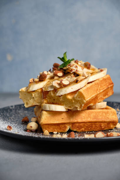 istock Sweet waffle with caramel and bananas sprinkled with hazelnuts and icing sugar on a white background 1204148520