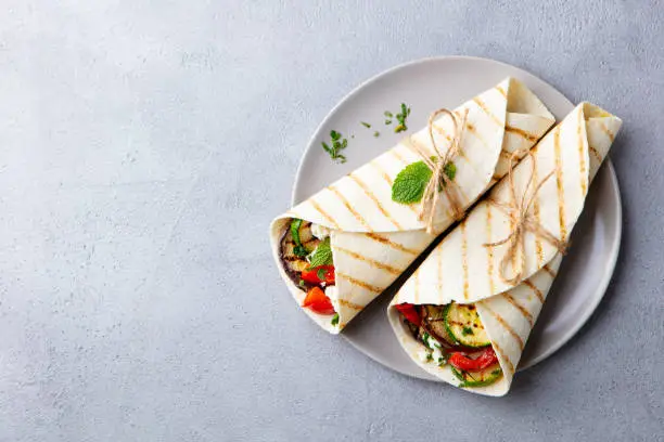 Photo of Wrap sandwich with grilled vegetables and feta cheese on a plate. Grey background. Copy space. Top view.
