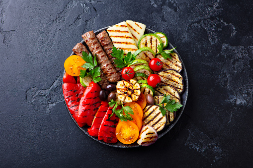 Grilled meat kebabs and vegetables on a black plate. Black stone background. Top view.