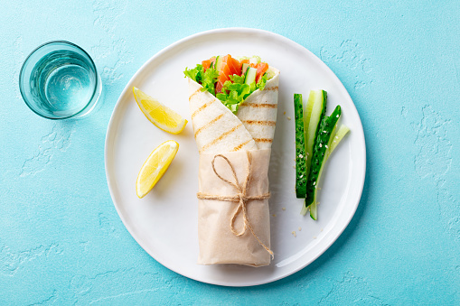 Wrap sandwich, roll with salmon, cucumber, salad on a white plate. Blue background. Top view.