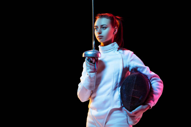 Teen girl in fencing costume with sword in hand isolated on black background Confident. Teen girl in fencing costume with sword in hand isolated on black background, neon light. Young model practicing and training in motion, action. Copyspace. Sport, youth, healthy lifestyle. fencing sport stock pictures, royalty-free photos & images