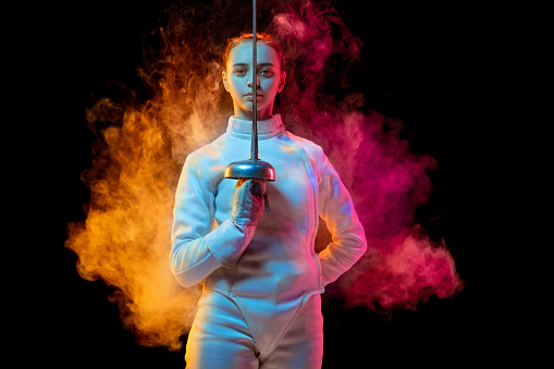 Historical time. Teen girl in fencing costume with sword in hand isolated on black background, neon lighted smoke. Practicing and training in motion, action. Copyspace. Sport, youth, healthy lifestyle.