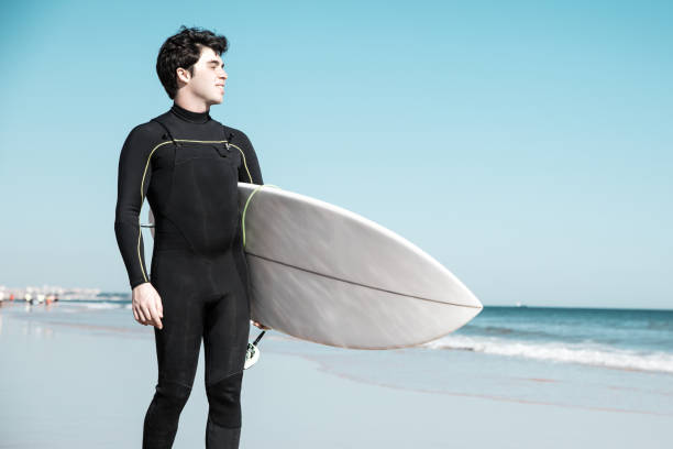 istock Smiling handsome young man holding surfboard on sunny beach 1204166371