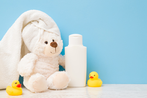 Smiling, lovely white teddy bear sitting. Towel on head. Yellow rubber ducks and shampoo bottle on shelf. Children bathing concept. Empty place for text on light blue wall. Pastel color. Closeup.