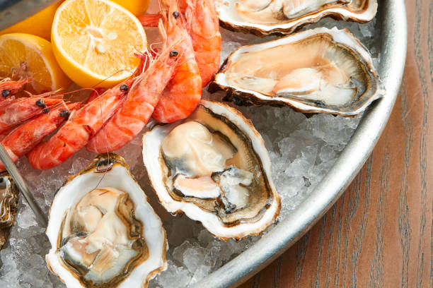 Appetizing assorted fresh seafood. Juicy oysters and boiled shrimps served on a metal dish on ice on a wooden background. Film effect during post. Soft focus stock photo