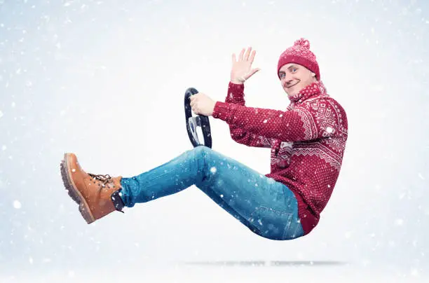 Happy smiling man in a red sweater, hat and scarf drives a car sends greetings to the camera, in snow. Auto driver concept