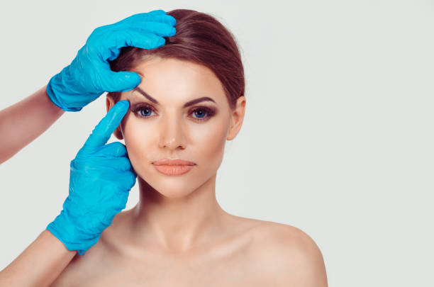 Upper eyelid blepharoplasty. Beautiful middle age woman getting ready for eyelid lift plastic surgery doctor hands in blue gloves point fingers to her eye on white. Beauty, people and health concept stock photo