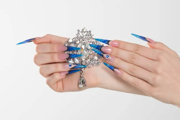 Nail Polish. Art Manicure. Modern style blue Nail Polish. Beauty hands holding white crystals gem stones diamonds broach showing its beauty. Stylish Colorful stiletto Nails isolated white background